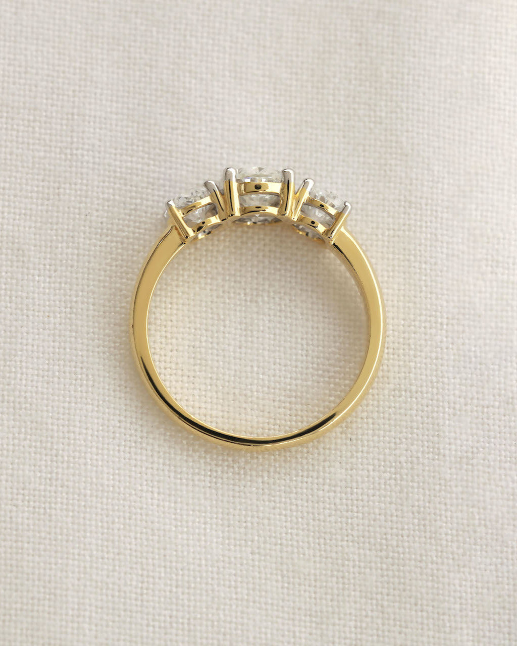 A gold three stone ring viewed from the side on the light colored background.