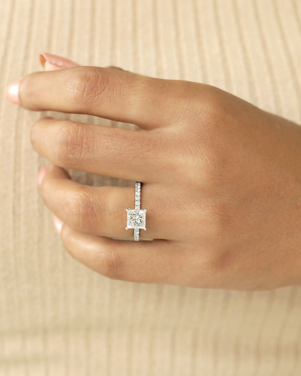 Close up shot of hand wearing a princess cut diamond ring on a beige background.