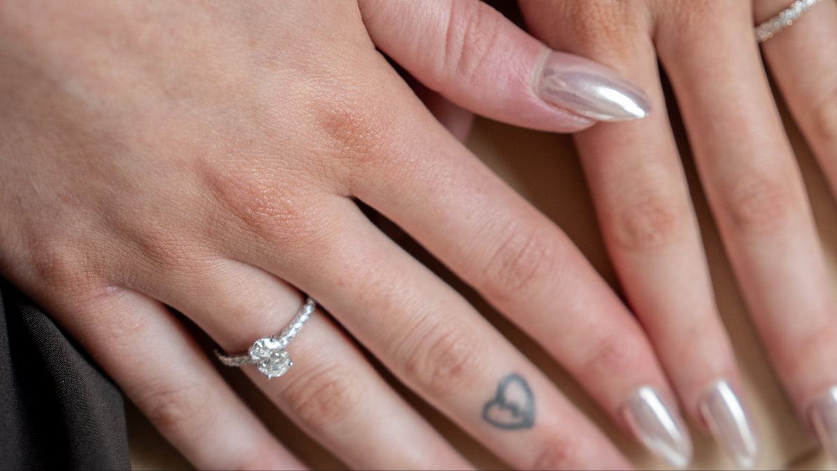 Close-up of two hands resting on top of each other, each wearing a silver ring. One hand has a small heart tattoo on the ring finger, and both nails are manicured in a metallic silver shade.