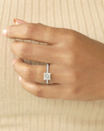 Close up shot of hand wearing a princess cut diamond ring on a beige background.