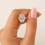 A close-up of a hand with light pink manicured nails holding a gold ring featuring an oval-cut clear gemstone, set against a soft beige background.