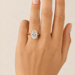 An oval-cut diamond engagement ring with a yellow gold band, shown worn on a hand against a light beige background.