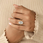 A close-up of a person's hands wearing a gold ring with a large oval-cut clear gemstone, set against a soft beige ribbed sweater background.