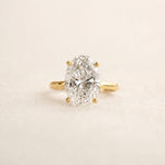 An oval-cut diamond ring with a yellow gold band, photographed on a clean, light beige textured background.