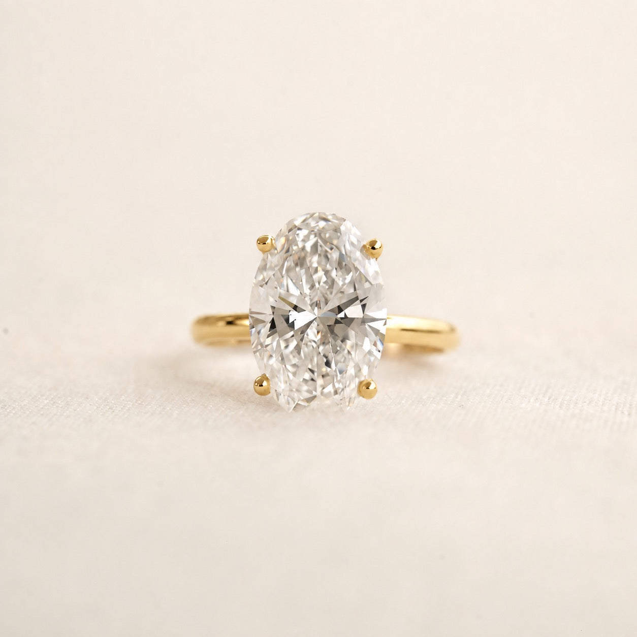 An oval-cut diamond ring with a yellow gold band, photographed on a clean, light beige textured background.