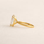 A yellow gold ring with a clear rectangular gemstone in a prong setting, shown from a side profile on a light beige textured background.