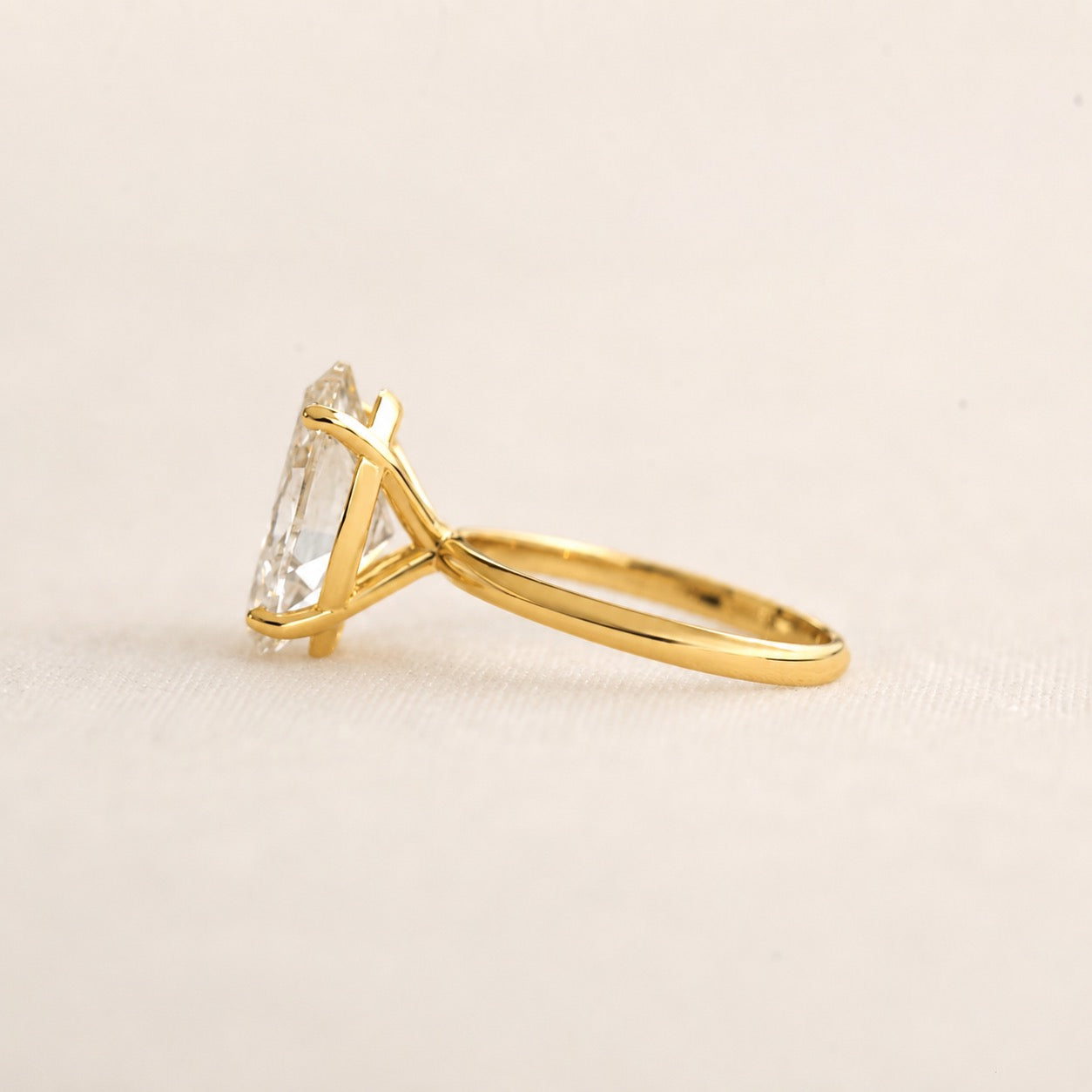 A yellow gold ring with a clear rectangular gemstone in a prong setting, shown from a side profile on a light beige textured background.