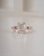 An engagement ring featuring a large cushion-cut diamond center stone in a prong setting, with a rose gold band accented by small round diamonds, set against a soft, warm beige background.