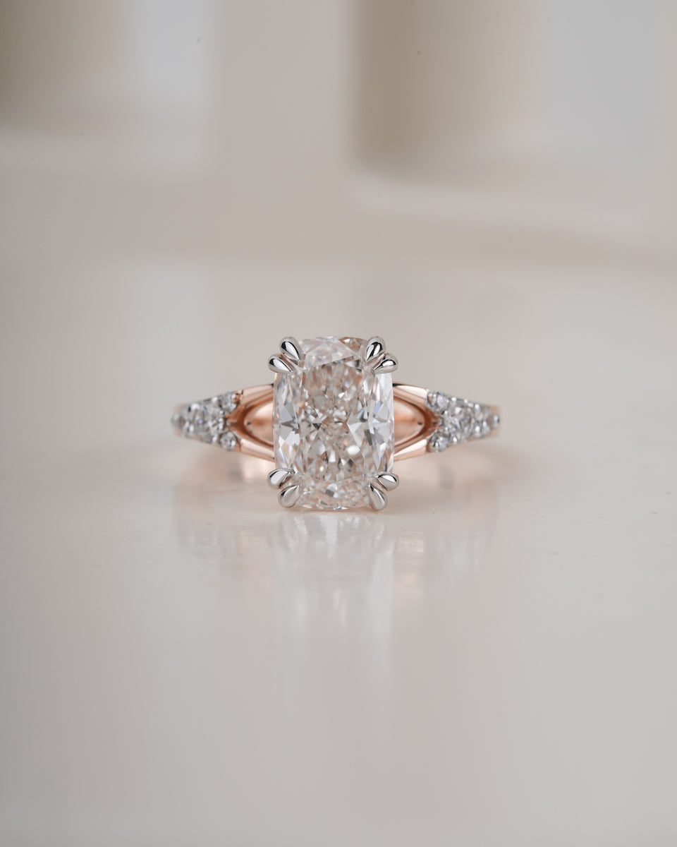 An engagement ring featuring a large cushion-cut diamond center stone in a prong setting, with a rose gold band accented by small round diamonds, set against a soft, warm beige background.