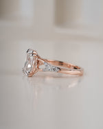A rose gold engagement ring featuring a large rectangular clear gemstone and small accent diamonds on the band, photographed on a soft, light beige background.