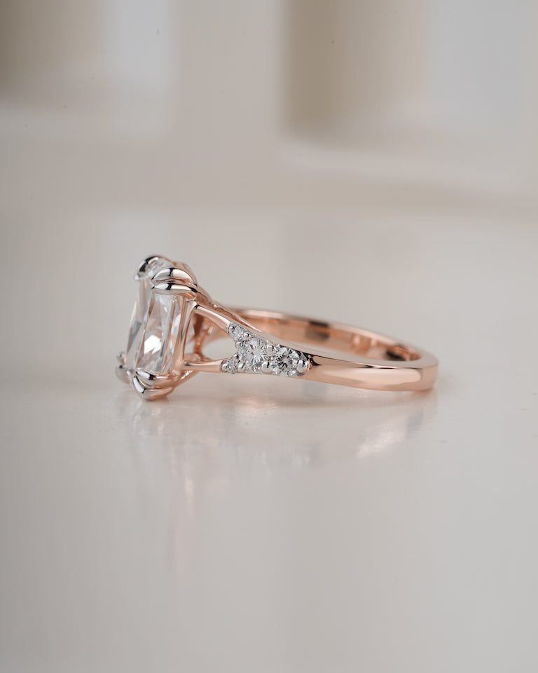 A rose gold engagement ring featuring a large rectangular clear gemstone and small accent diamonds on the band, photographed on a soft, light beige background.