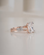 A rose gold engagement ring featuring a large rectangular clear gemstone and small accent diamonds on the band, set against a soft, warm off-white background.