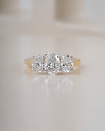 A three-stone diamond ring with round brilliant-cut diamonds set in a yellow gold band, resting on a light beige reflective surface.