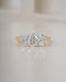 A three-stone diamond ring with round brilliant-cut diamonds set in a yellow gold band, resting on a light beige reflective surface.