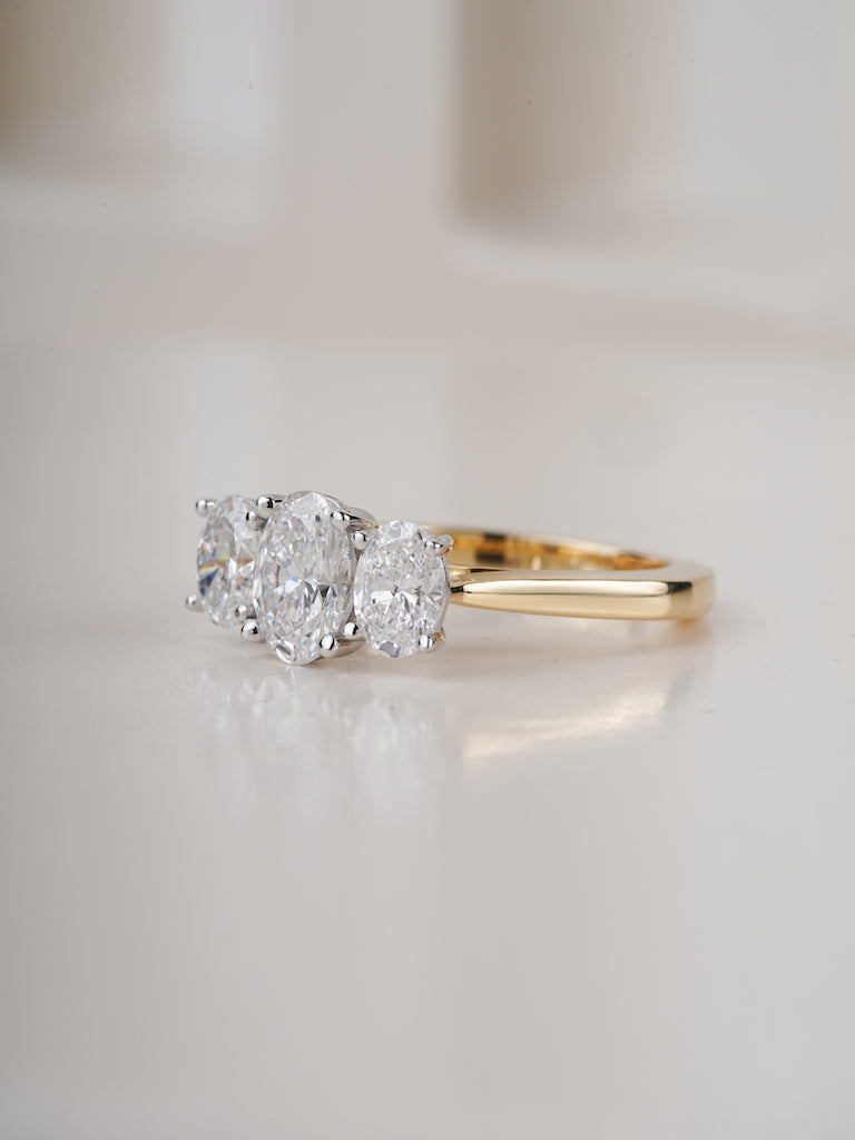 A yellow gold three-stone diamond ring with oval-cut center stone and round side stones, set on a soft white reflective surface.