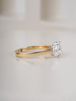 A classic solitaire engagement ring featuring a round brilliant-cut clear gemstone in a four-prong setting on a polished yellow gold band, photographed on a clean, light beige reflective surface.