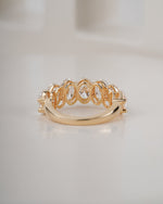 A gold ring featuring a series of oval-cut clear gemstones in an openwork pattern, set on a polished gold band, photographed on a light beige surface.