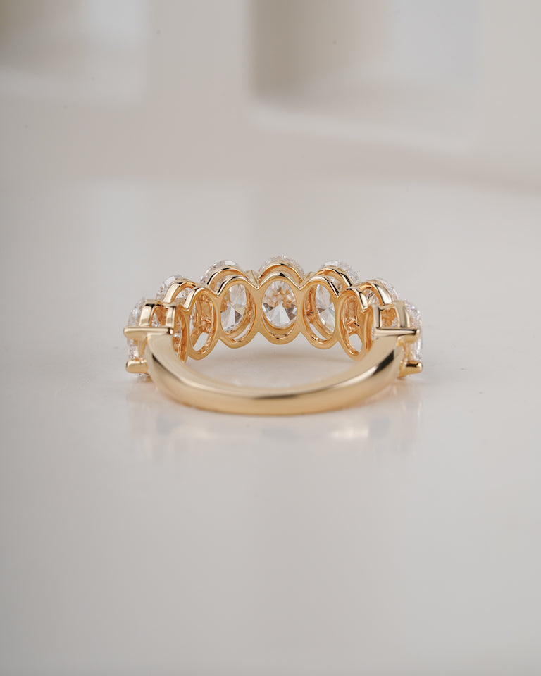 A gold ring featuring a series of oval-cut clear gemstones in an openwork pattern, set on a polished gold band, photographed on a light beige surface.