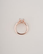 A rose gold ring with a heart-shaped clear gemstone in a four-prong setting, shown from a top-down view on a light gray background.