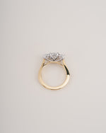 A gold ring with a three-stone cluster setting featuring clear gemstones, photographed from a top-down angle on a light beige background.