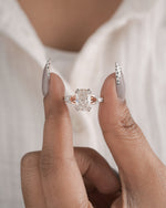 A close-up of a person's hands holding a rose gold engagement ring with a marquise-cut center stone and small side stones, set against a soft white shirt background.