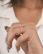 A close-up of a person's hand wearing a rose gold ring with a clear marquise-cut gemstone and small accent stones, set against a soft white shirt background.