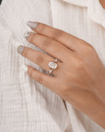 A close-up of a hand wearing a rose gold ring with a cushion-cut clear gemstone, resting on a textured white fabric background.