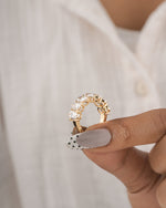 A gold ring with clear gemstones held between fingers against a soft white shirt background.
