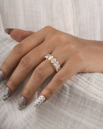 A close-up of a hand wearing a gold band ring set with clear round gemstones, resting on a textured white fabric background.