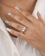 A close-up of a hand wearing a gold ring with three clear round-cut gemstones, styled with silver and white nail art over a white textured top.