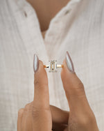 A close-up of a person's hands with long silver manicured nails holding a gold ring with a rectangular emerald-cut clear gemstone, set against a soft-focus white textured shirt.