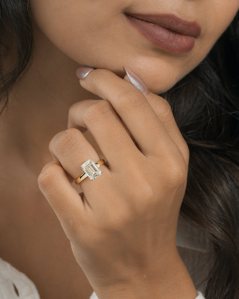 A close-up of a woman's hand wearing a yellow gold ring with an emerald-cut clear gemstone, set against her face and dark hair.