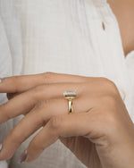 A gold ring with a rectangular clear gemstone worn on a person's finger, shown against a soft white textured blouse background.