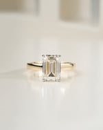 An emerald-cut diamond ring with a yellow gold band, centered on a clean, light-colored reflective surface.