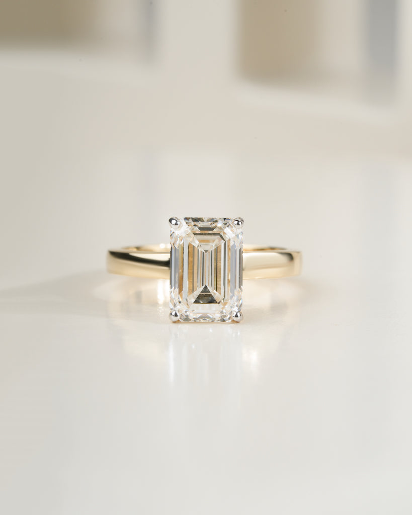 An emerald-cut diamond ring with a yellow gold band, centered on a clean, light-colored reflective surface.