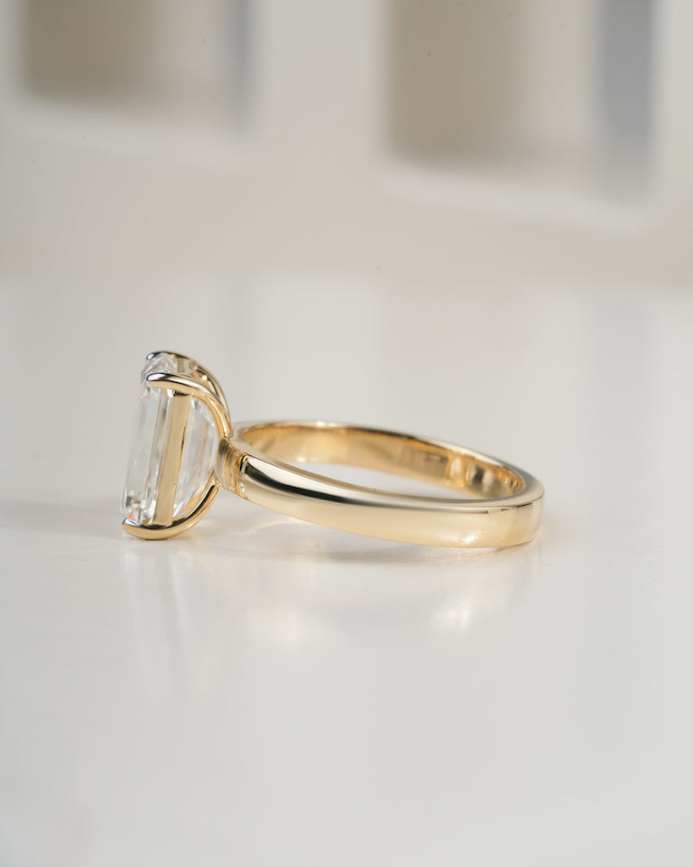 A yellow gold ring with a large rectangular clear baguette-cut gemstone, shown in a side profile on a reflective white surface.