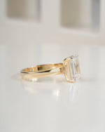 A gold ring with a large rectangular clear gemstone, photographed on a clean white surface with a softly blurred background.