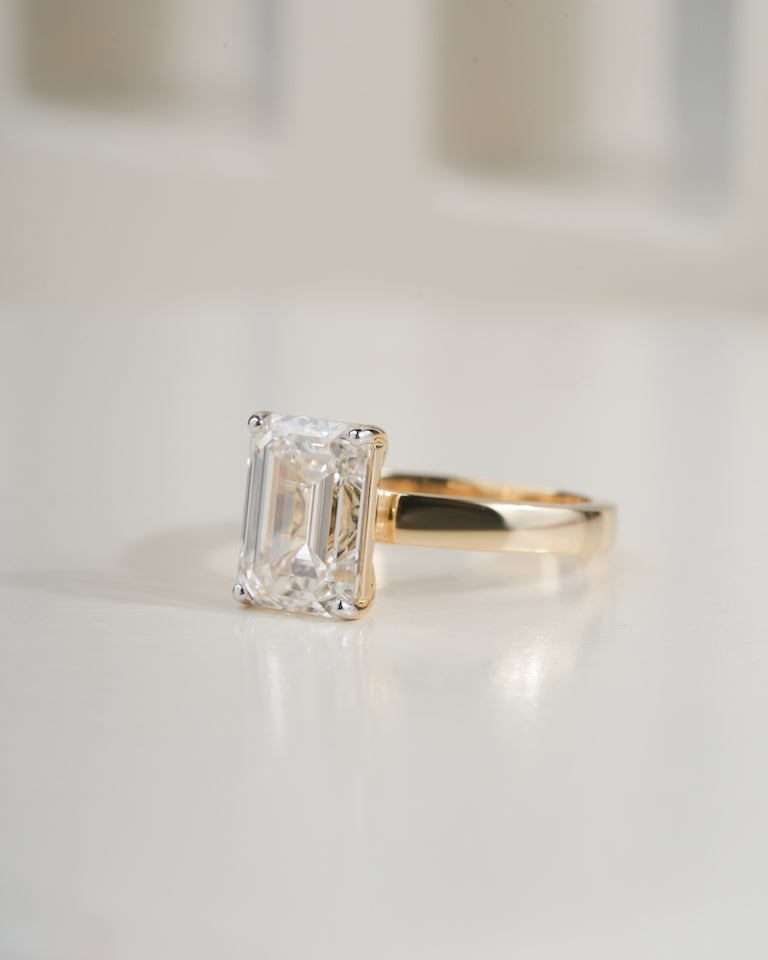 An emerald-cut diamond ring set in a yellow gold band, resting on a clean white reflective surface.
