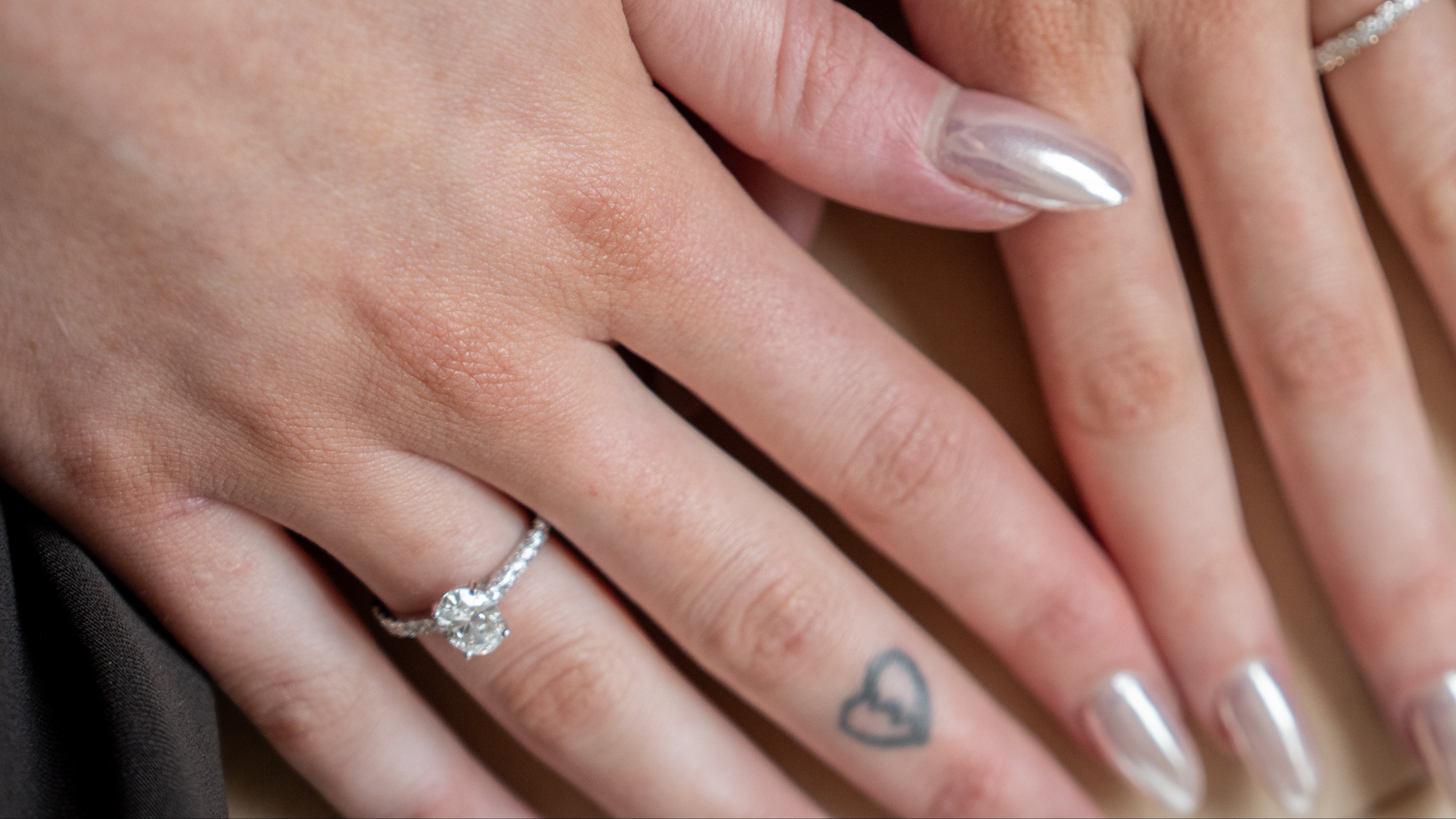 Close-up of two hands resting on top of each other, each wearing a silver ring. One hand has a small heart tattoo on the ring finger, and both nails are manicured in a metallic silver shade.