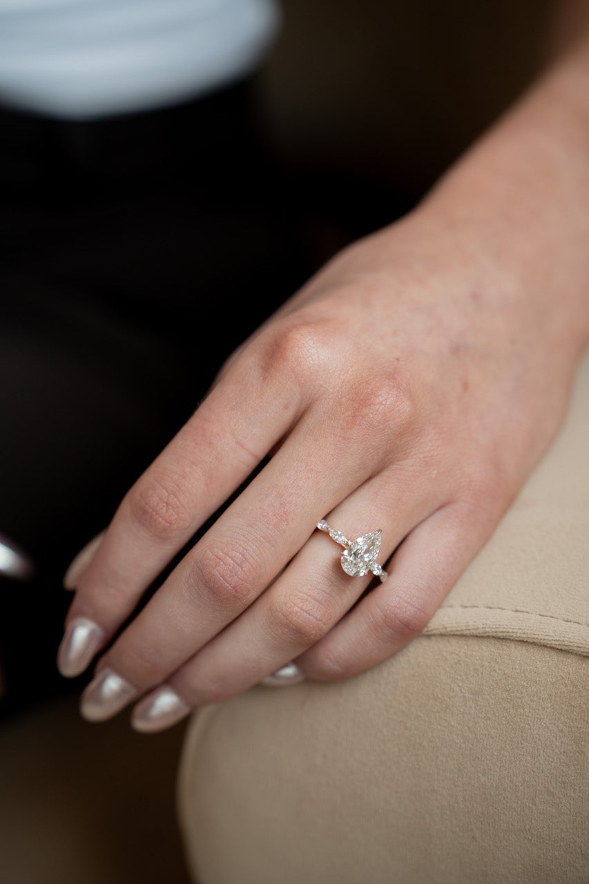 A close-up of a hand wearing a silver-toned ring with a floral cluster of clear gemstones, resting on a beige surface.